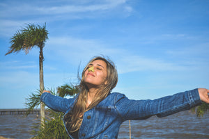 Young, blonde women in denim jacket opening arms looking up into clear sky with eyes closed